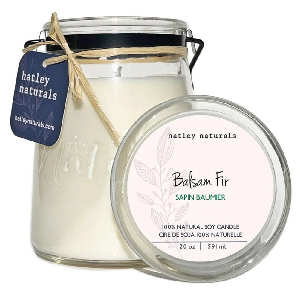 A glass jar candle with white wax, a black metal handle, and a twine-tied hanging tag reading hatley naturals, shown alongside its round lid label that says Balsam Fir / Sapin Balmer on a white background.