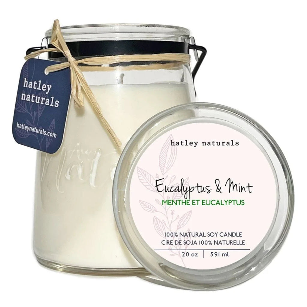 A glass jar candle with white wax, a black metal lid, and a twine tag reading hatley naturals, shown next to its round lid label that says Eucalyptus & Mint on a white background.